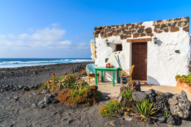 Ein weißes Haus mit grünen Tisch und Stühlen an einem Strand mit schwarzem Sand.