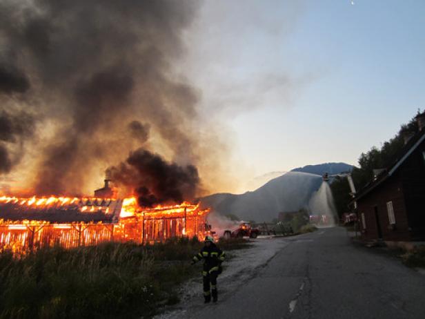 Ein brennendes Gebäude mit einem Feuerwehrmann im Vordergrund und Löscharbeiten im Hintergrund.