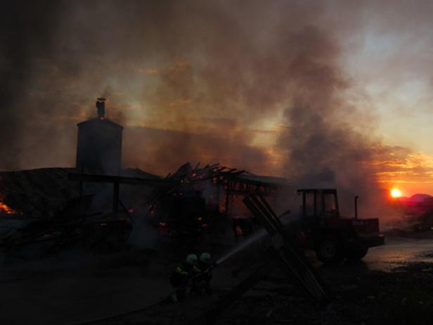 Feuerwehrleute bekämpfen einen Brand in einem landwirtschaftlichen Gebäude bei Sonnenuntergang.