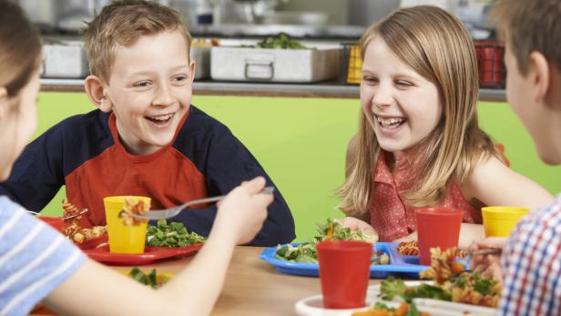 Kinder sitzen lachend beim Mittagessen in der Schulkantine.