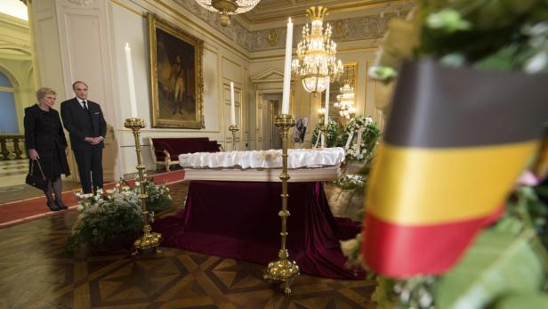 In einem prunkvollen Raum steht ein Sarg, umgeben von Kerzen und Blumen, mit der belgischen Flagge im Vordergrund.