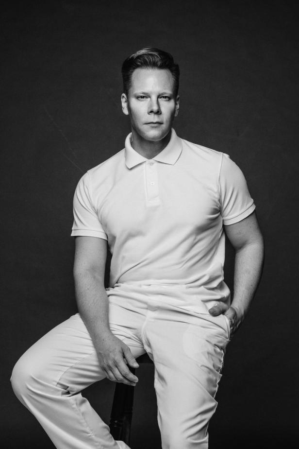 Ein Mann in weißem Polohemd und Hose sitzt auf einem Stuhl vor einem dunklen Hintergrund.