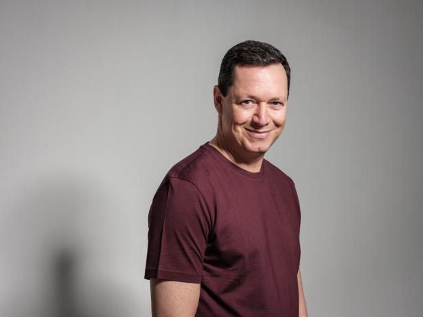 Ein lächelnder Mann mit einem weinroten T-Shirt vor einem grauen Hintergrund.