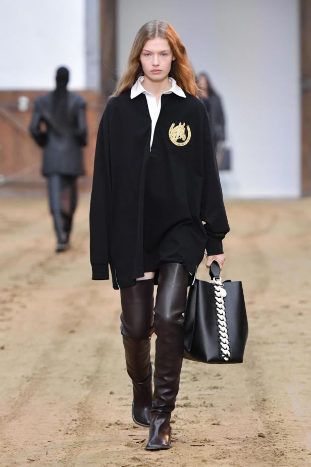 Ein Model auf dem Laufsteg trägt ein schwarzes Kleid mit Pferdeemblem und braune Stiefel.