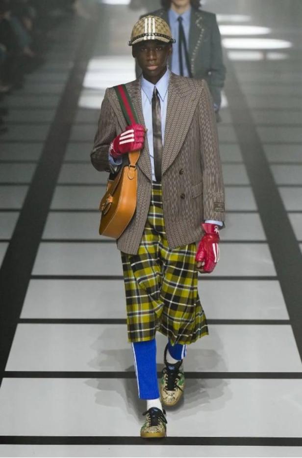 Ein dunkelhäutiges Model auf dem Laufsteg, gekleidet in einen karierten Rock, Blazer und eine Kappe mit dem Gucci-Logo.