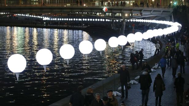 Eine Reihe leuchtender Kugeln erstreckt sich über einen Fluss, während Menschen am Ufer entlanggehen.