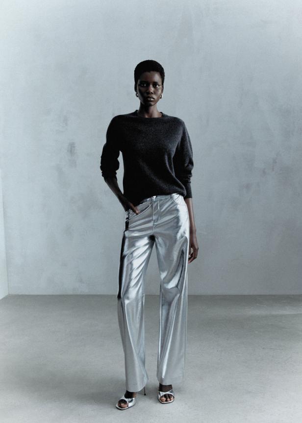 Eine Frau mit dunkler Haut trägt einen grauen Pullover und eine glänzende silberne Hose.