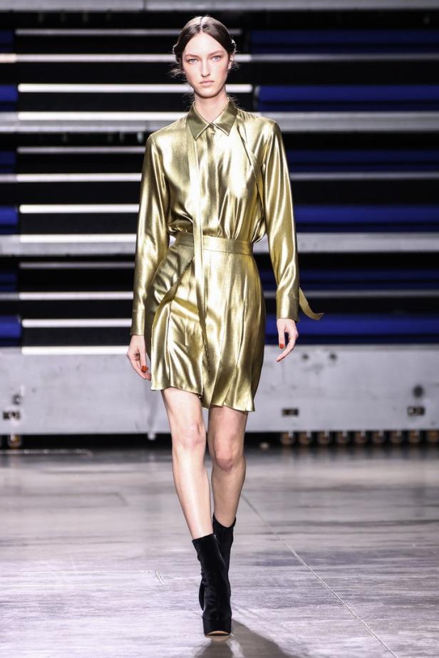 Eine Frau in einem goldenen Hemdkleid und schwarzen Stiefeln auf einem Laufsteg.