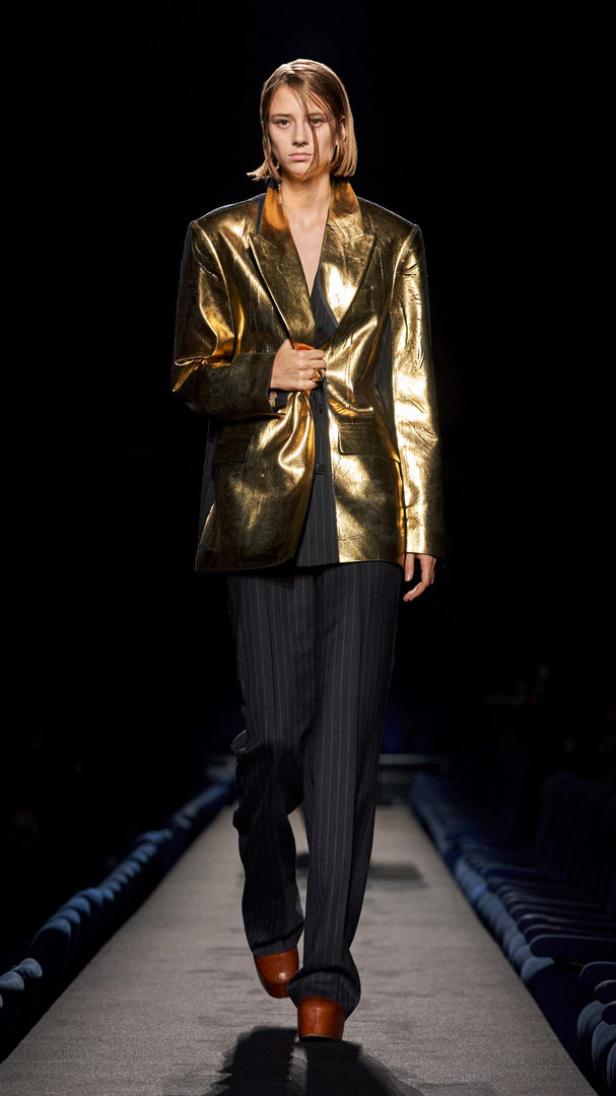 Eine Frau in einem goldenen Blazer und einer gestreiften Hose auf einem Laufsteg.