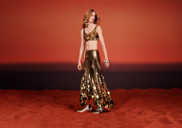 Eine Frau in einem goldenen Pailletten-Top und Rock vor einem orangefarbenen Hintergrund.