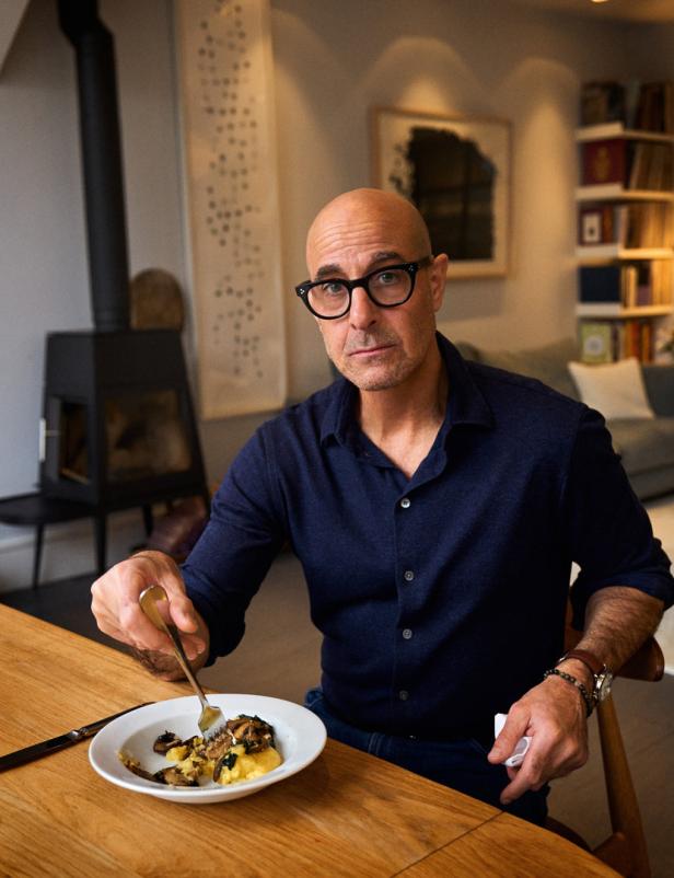 Stanley Tucci sitzt an einem Holztisch und isst ein Gericht mit Pilzen.