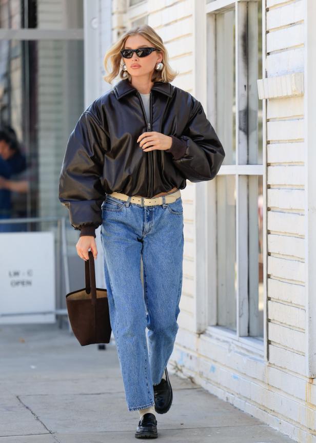 Eine Frau mit Sonnenbrille, Lederjacke und Jeans geht die Straße entlang.