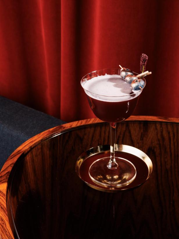 Ein Cocktail, garniert mit Blaubeeren, steht auf einem dunklen Holztisch.