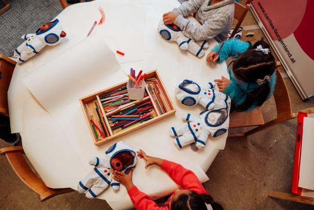 Kinder sitzen an einem Tisch und bemalen Papier und Astronauten-Stofftiere.