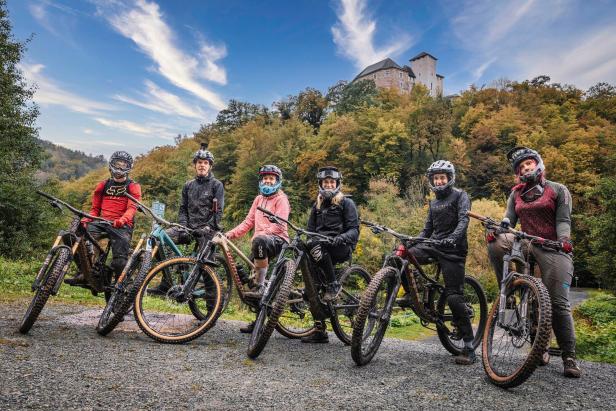 Eine Gruppe Mountainbiker posiert mit ihren Fahrrädern vor einer Burg auf einem Hügel.