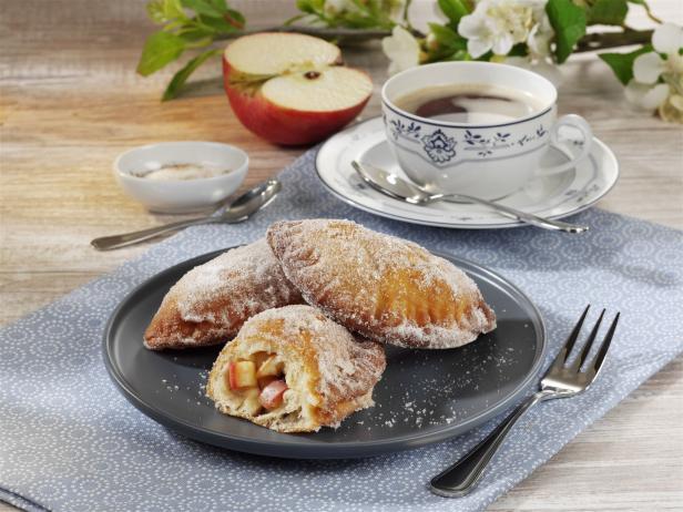 Apfeltaschen mit Puderzucker auf einem Teller, dazu eine Tasse Kaffee und eine halbe Apfel.