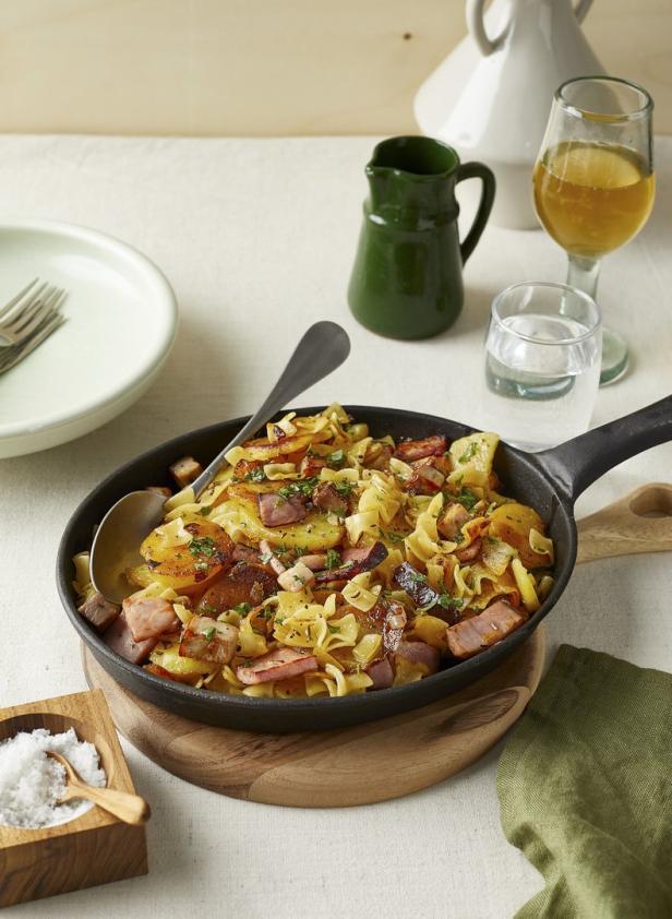 Eine Pfanne mit Kartoffeln, Schinkenwürfeln und Bandnudeln steht auf einem Tisch.