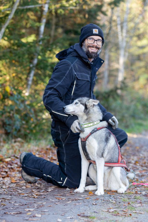 Ein Mann kniet neben einem sitzenden Husky auf einem Waldweg.