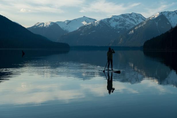 Eine Person steht auf einem Paddleboard auf einem ruhigen See vor einer Bergkulisse.