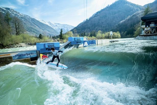 Ein Surfer reitet auf einer Welle im „The River Wave“ Park vor einer Bergkulisse.