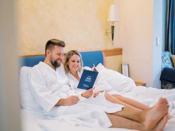 Ein Paar im Bademantel liegt im Bett und liest eine Broschüre.