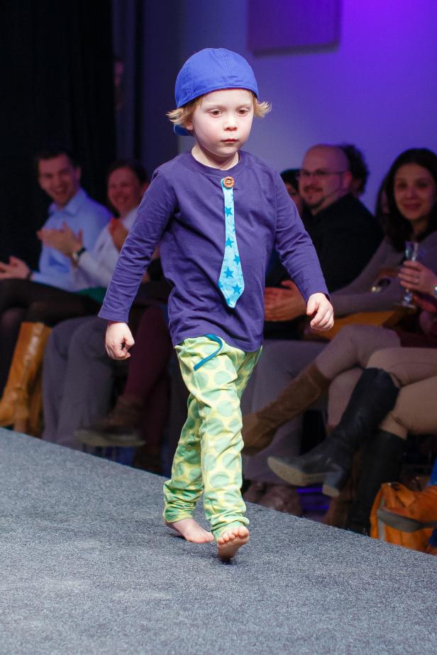 Ein barfüßiger Junge mit Kappe und Sternenkrawatte läuft auf einem Laufsteg.