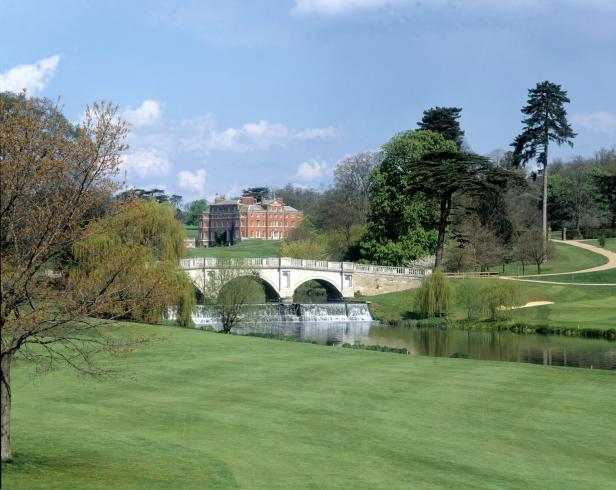 Ein Herrenhaus aus rotem Backstein überblickt einen Golfplatz mit einem Fluss und einer Steinbrücke.