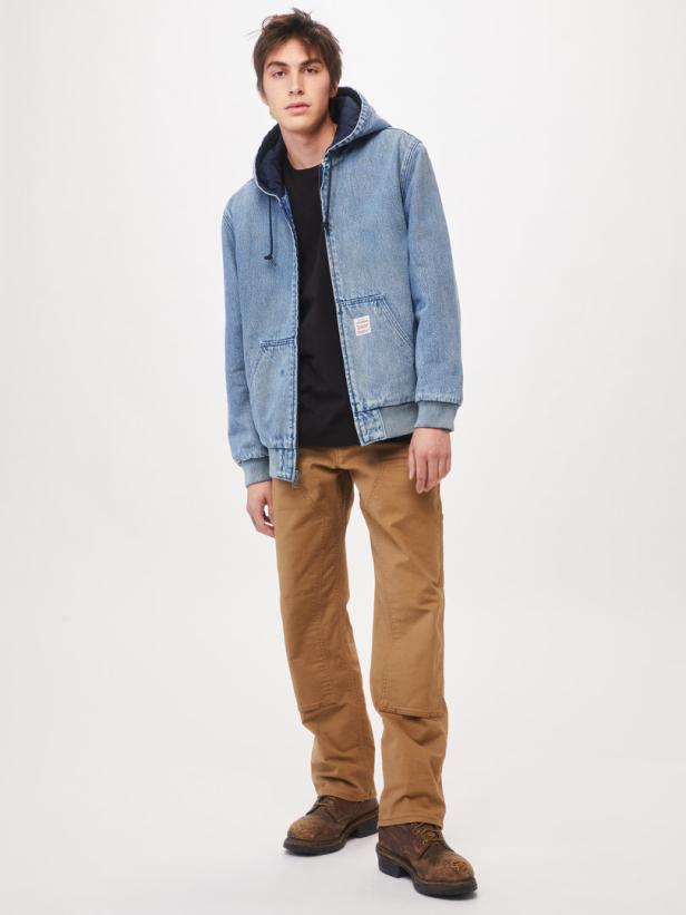 Ein Mann mit Jeansjacke, braunen Hosen und Stiefeln steht vor einem weißen Hintergrund.