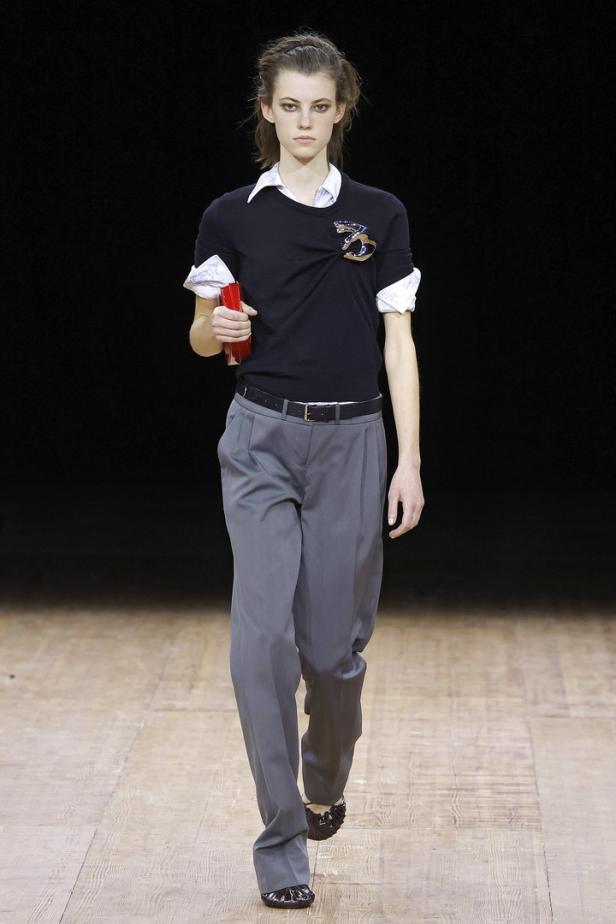 Ein Model auf dem Laufsteg, gekleidet in eine graue Hose und ein schwarzes T-Shirt mit Applikation.