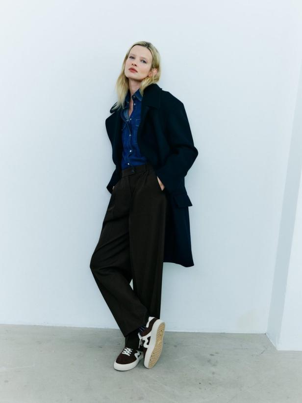Eine blonde Frau posiert in einem dunklen Mantel, einer Jeansbluse und braunen Hosen vor einer weißen Wand.
