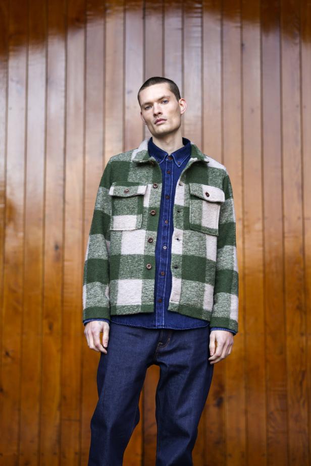 Ein Mann mit blauem Hemd und karierter Jacke steht vor einer Holzwand.