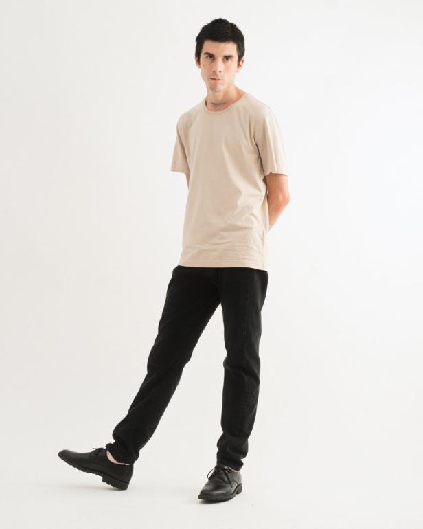 Ein Mann in beigem T-Shirt, schwarzen Jeans und Lederschuhen posiert vor einem weißen Hintergrund.