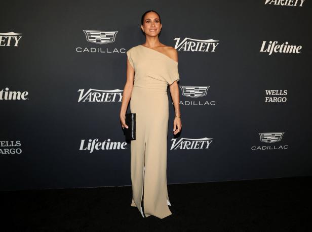 Meghan Markle in einem beigen Kleid auf einer Veranstaltung mit Sponsorenlogos im Hintergrund.