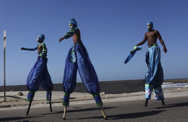 Drei Stelzenläufer in blauen Kostümen treten auf einer Straße am Meer auf.