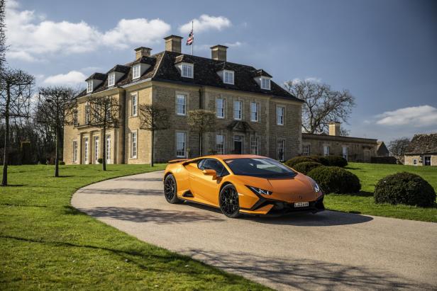 Ein orangefarbener Lamborghini steht vor einem großen Herrenhaus mit britischer Flagge.