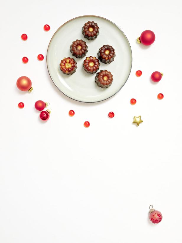 Canelés auf einem Teller, umgeben von roten Weihnachtskugeln und Dekorationen.