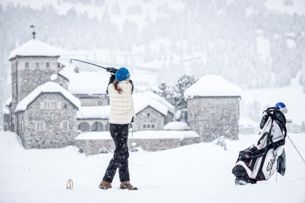 Eine Frau golft im Schnee vor einer verschneiten Häuserkulisse.