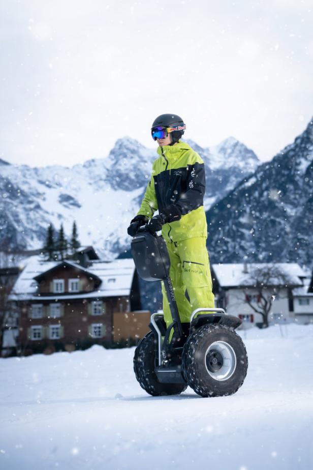 Eine Person in Skikleidung fährt mit einem Segway durch den Schnee vor einer Bergkulisse.