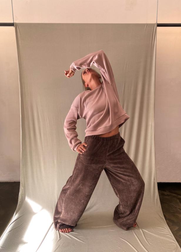 Eine Frau in Cordhose und Sweatshirt posiert vor einem neutralen Hintergrund.