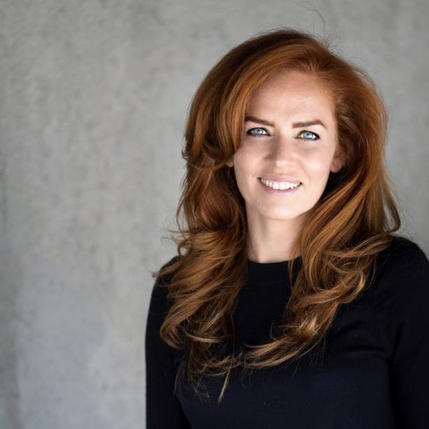 Eine lächelnde Frau mit roten Haaren und blauen Augen vor einem grauen Hintergrund.