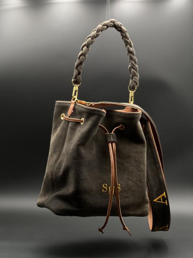 Eine braune Wildledertasche mit geflochtenem Griff und goldenen Initialen vor einem dunklen Hintergrund.