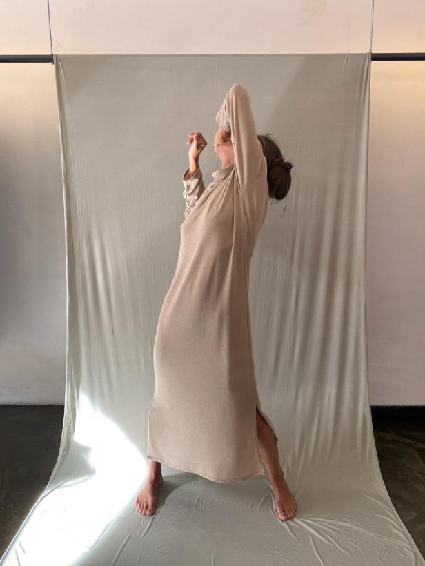 Eine Frau in einem langen, beigen Kleid posiert vor einem hellen Hintergrund.
