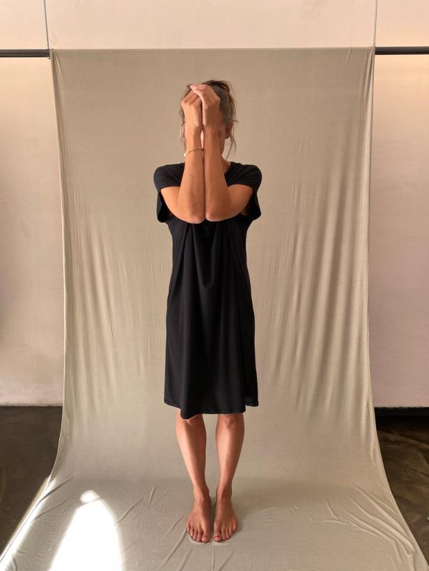 Eine Frau in einem schwarzen Kleid steht barfuß vor einem neutralen Hintergrund und bedeckt ihr Gesicht mit den Händen.