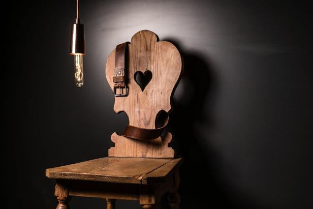 Ein Holzstuhl mit Herzmuster und ein Ledergürtel vor einer dunklen Wand.