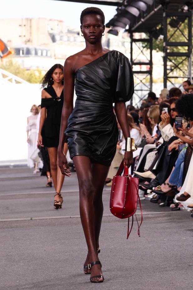 Eine Frau in einem schwarzen, schulterfreien Kleid mit roter Handtasche auf einem Laufsteg.