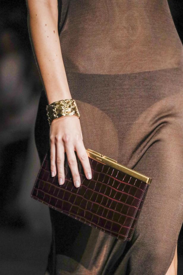 Eine Frau trägt ein goldenes Armband und eine burgunderfarbene Tom-Ford-Clutch.