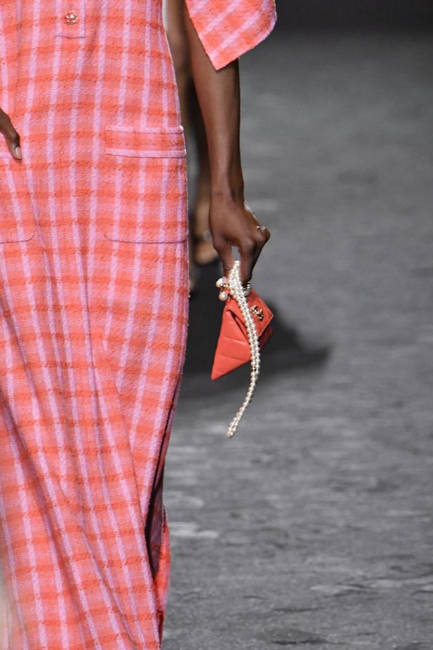 Ein Model trägt ein kariertes Kleid und eine orangefarbene Chanel-Tasche mit Perlenkette.