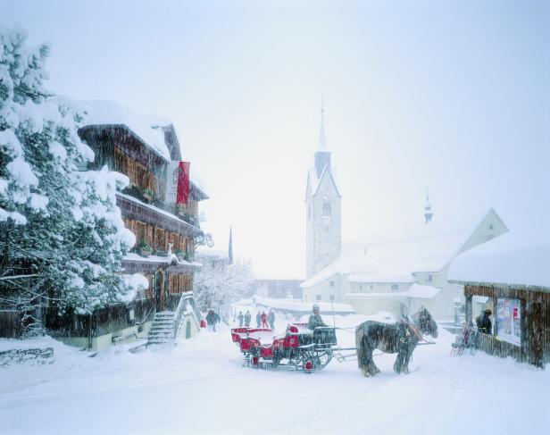 Eine Pferdekutsche fährt durch ein verschneites Dorf mit einer Kirche im Hintergrund.