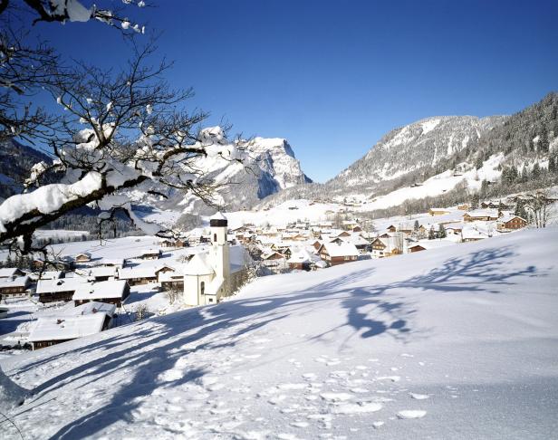 Ein verschneites Dorf mit einer Kirche vor einer Bergkulisse an einem sonnigen Tag.