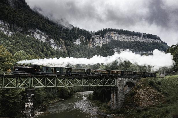 Eine Dampflokomotive fährt über eine grüne Stahlbrücke vor einer bewaldeten Bergkulisse.
